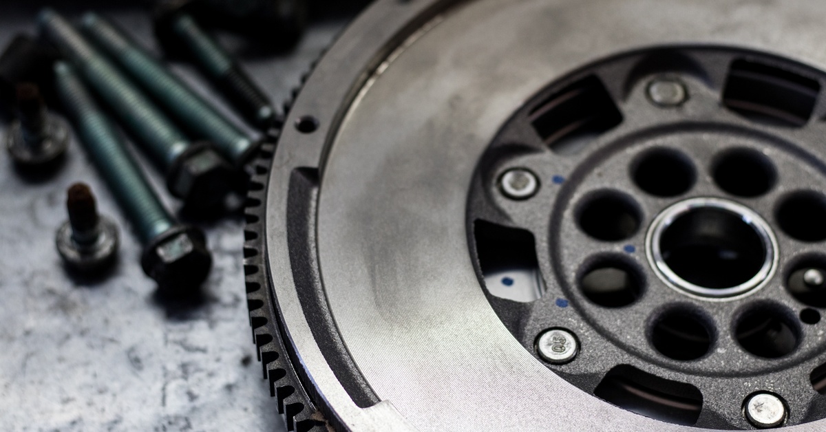 A new dual-mass flywheel on a metal workbench at close range. Lug nuts and long screws are beside the flywheel.