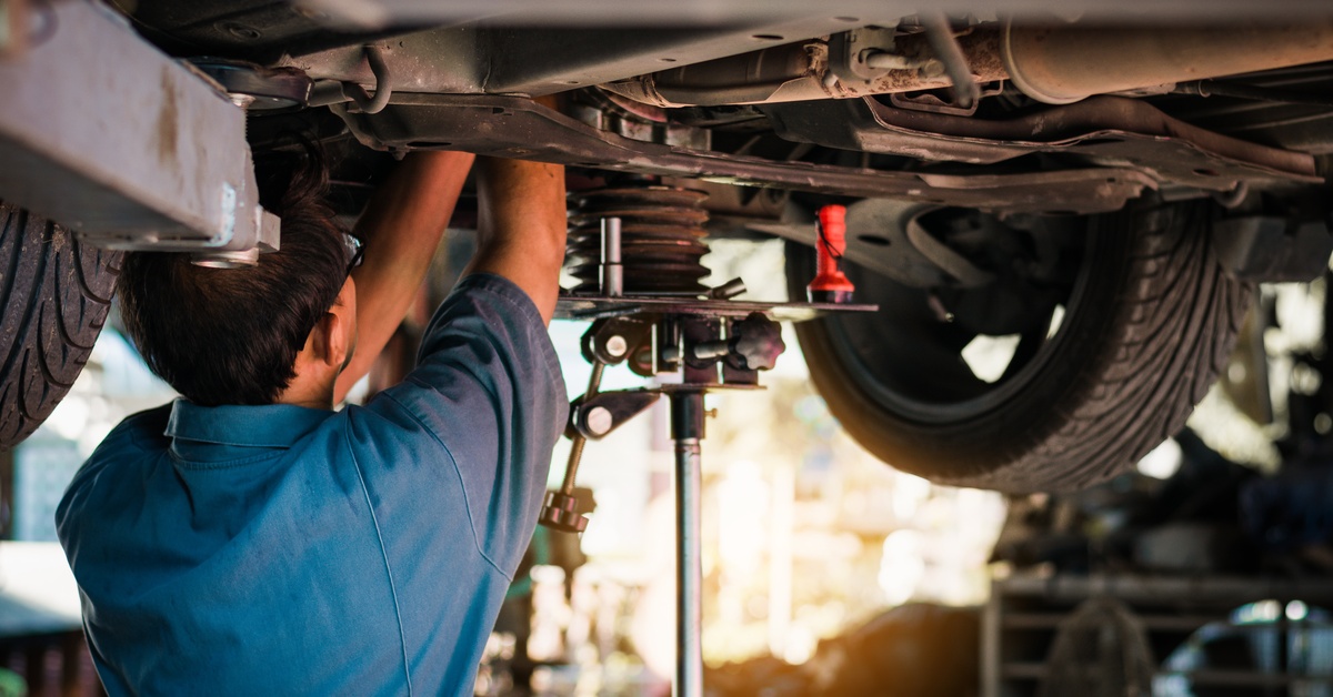 A rear view of a mechanic in a work shirt and glasses working on the suspension of a vehicle elevated on a lift.