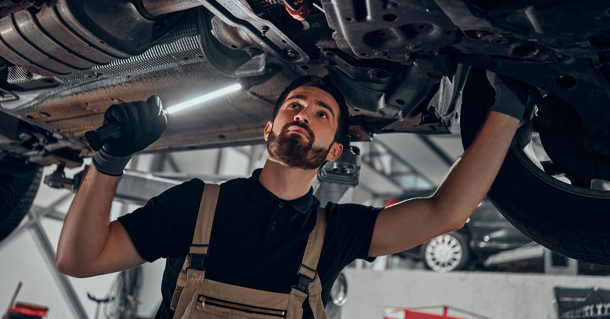 A mechanic in a black shirt and overalls with a light inspects the undercarriage of a vehicle elevated on a lift.