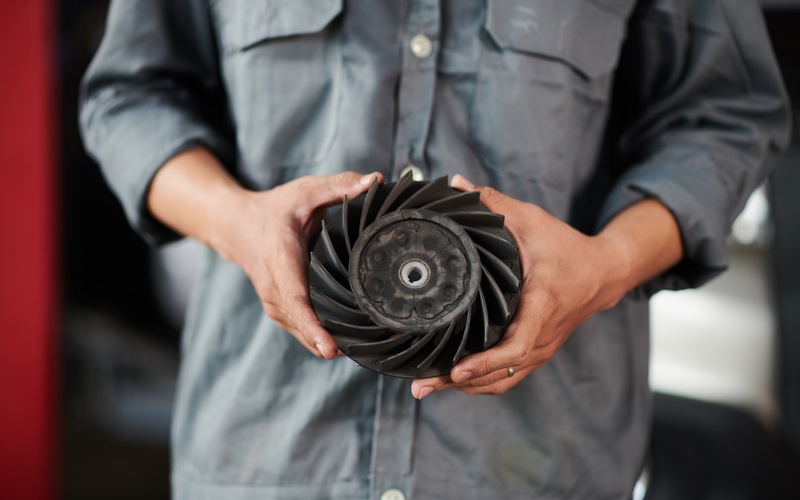 An automotive mechanic in a gray work shirt holds a used engine cooling fan for a car clutch between his hands.