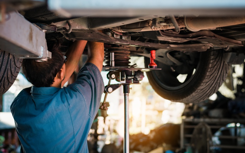 A worker in a blue shirt and safety glasses works on the undercarriage of a vehicle on a hydraulic lift.