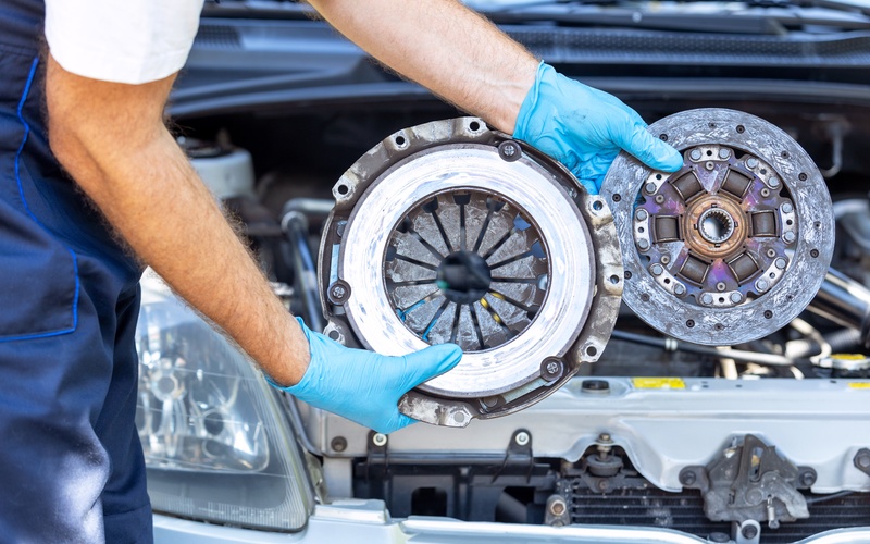 An automotive technician in blue overalls holds a used car pressure plate and clutch disc in front of a vehicle.
