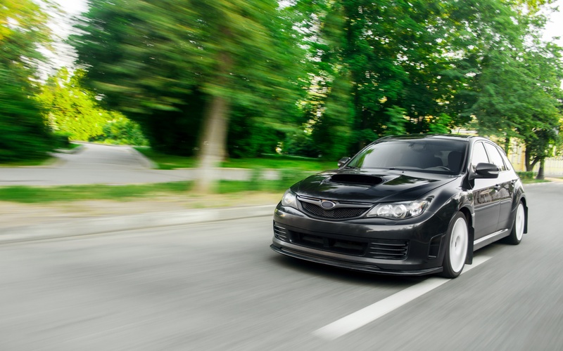 A front view of a black Subaru sedan driving on a road with trees, grass, and another road blurred behind it.