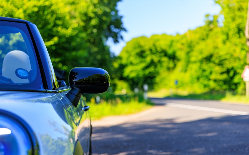 A close-up of the front and side of a new white Mini Cooper parked beside a road surrounded by green trees.
