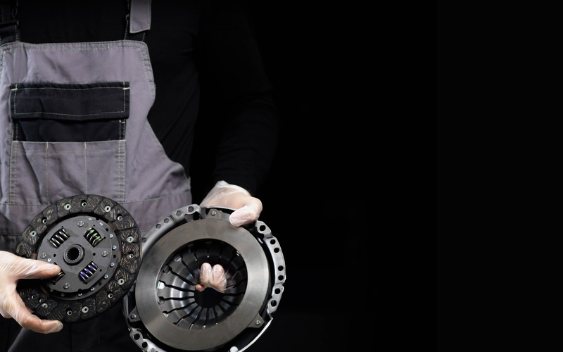 A close-up of a mechanic in gray overalls and a black shirt holding a brand-new clutch kit with transparent latex gloves.