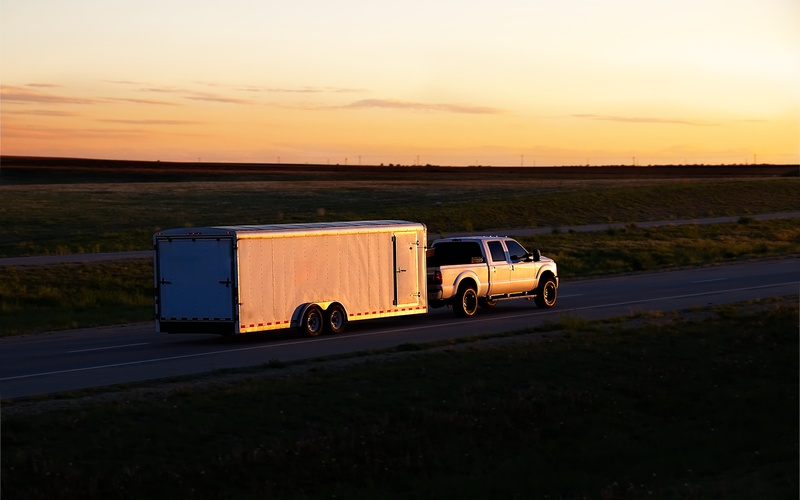 A pickup truck driving away from the viewer and towing a white box trailer down an empty rural highway during sunset hours.