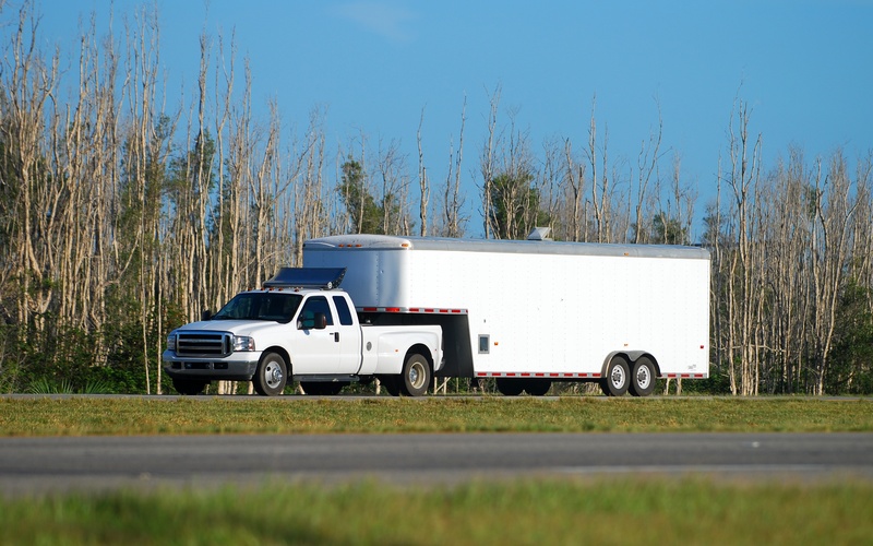 A white heavy-duty pickup truck towing a large, white fifth-wheel box trailer on an empty road during the day.