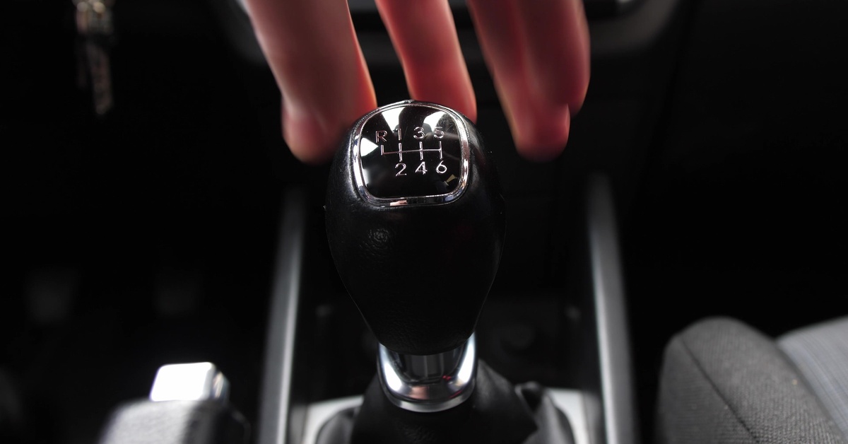The manual transmission gear stick of a car, showing six gears and a reverse gear, as fingers move to grasp it.