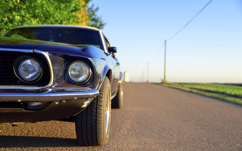 The front end grille and headlight of a blue, classic Ford Mustang on an empty rural road with silos in the background.