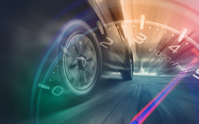 A close-up of the wheels of a car spinning in a blur, with a transparent overlay of the rpm gauge and dial of the car.