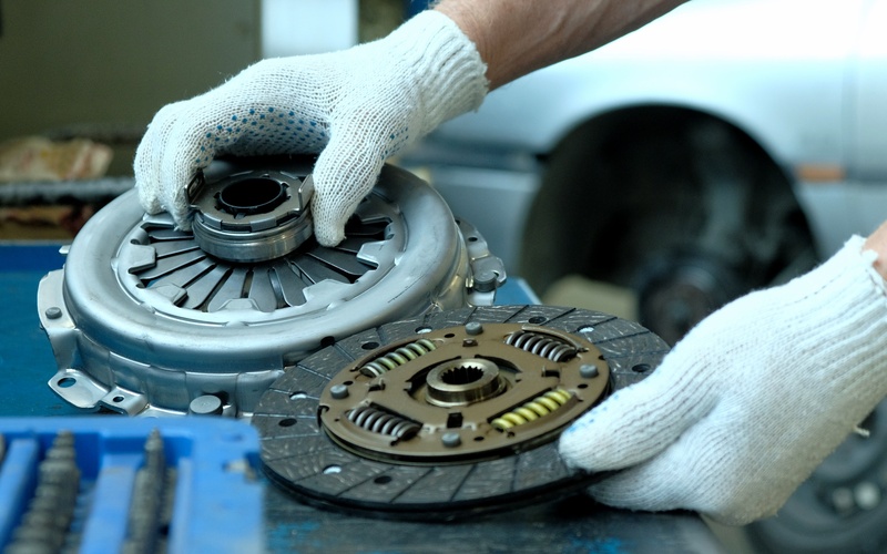 A close-up of used automotive clutch kit components. Two hands with white gloves hold the clutch disc and exhaust bearing.