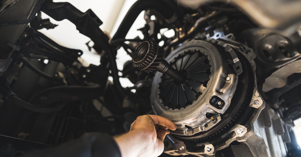 A close-up view looking up at a mechanic reaching his hand to install a new clutch kit in an elevated car.