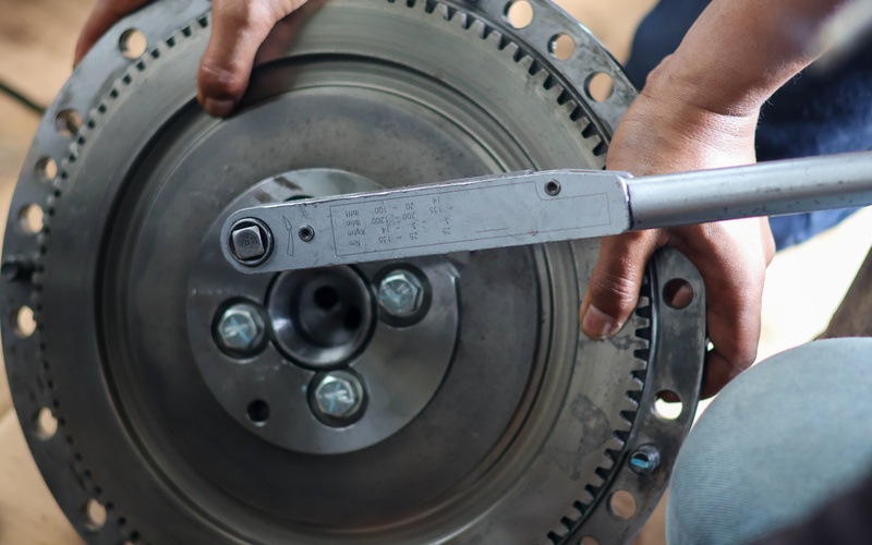 A close-up of a metal flywheel behing held by two hands while someon else uses a torque wrench to tighten its bolts.