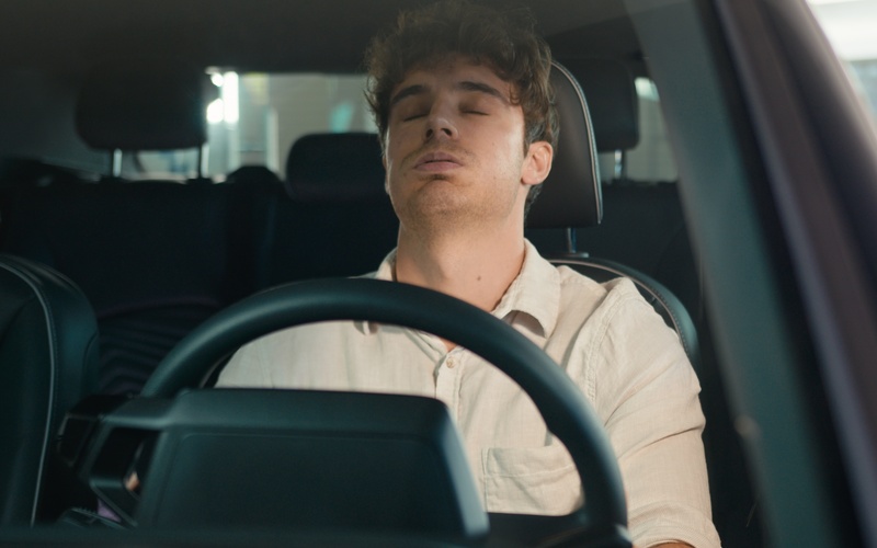A view through the windshield of a man in a white shirt exhaling in frustration behind the wheel of a car.