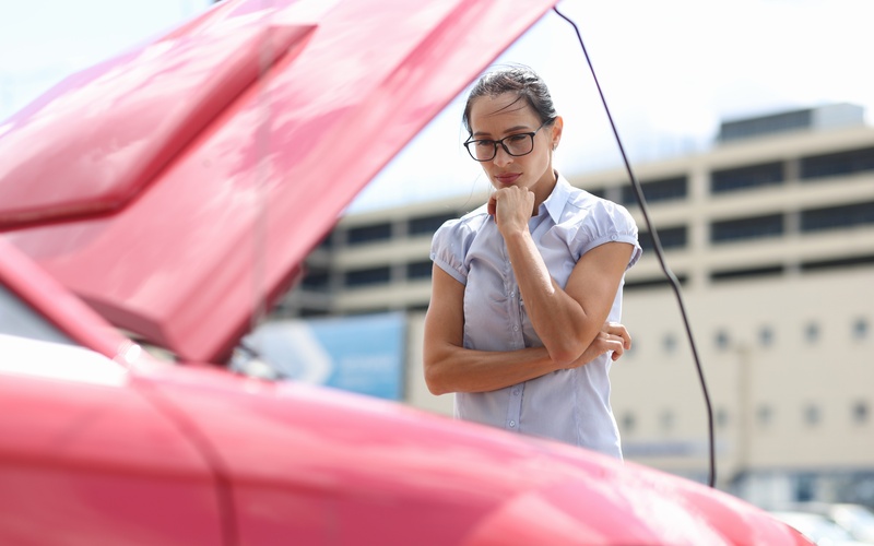 A woman in glasses and blue blouse looks at the engine of her car with her hood propped open on a sunny day.