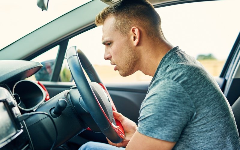 A young man in a grey t-shirt in the driver's seat of a modern car examines the dashboard with a confused look.