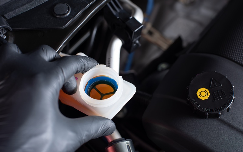 A close-up of a hand with a black latex glove examining the opening of a car's brake fluid reservoir.