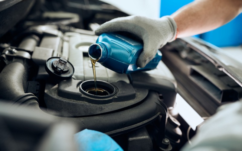 A close-up of a man's hands with gloves holding a blue bottle over the opening of a car to pour amber liquid down.
