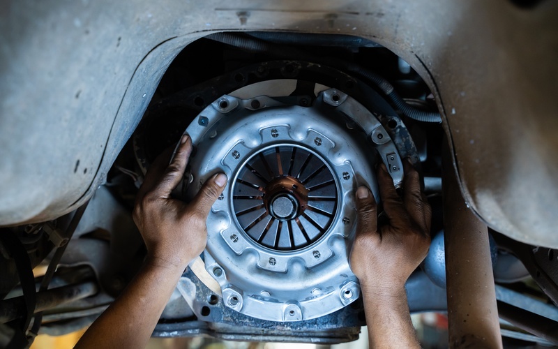 A close-up of the clutch plate and system of a car elevated on a lift as a person holds the cover with their hands.