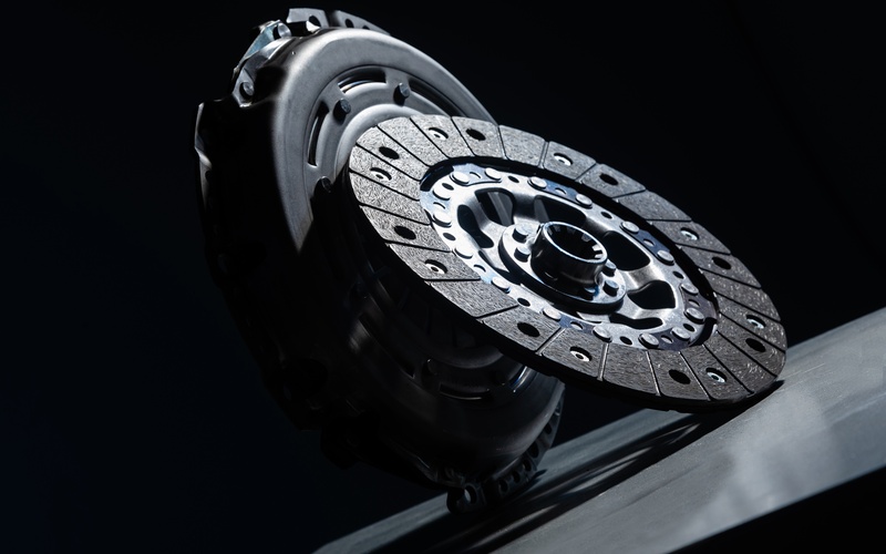 A close-up of a clutch disc leaning against the clutch basket on a grey table with a black background.