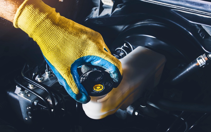 A close-up of a hand with a yellow and blue glove removing the cap from a car's brake fluid reservoir.