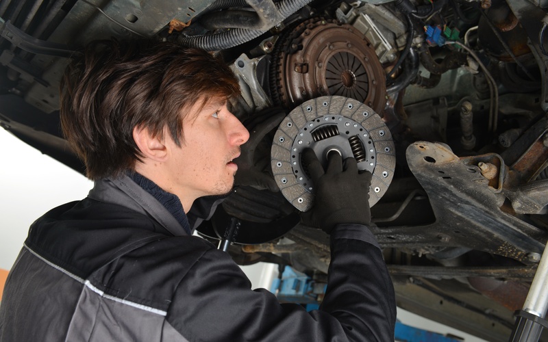 A young mechanic with black gloves and jumpsuit installing a clutch disc into the undercarriage of an elevated car.