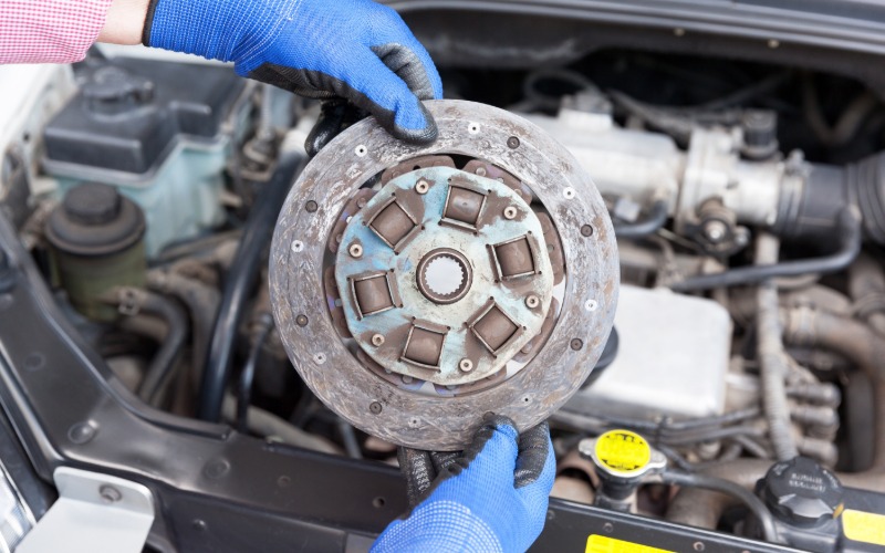 A close-up of a pair of hands with blue gloves holding an old car clutch plast disc over an open engine hood.