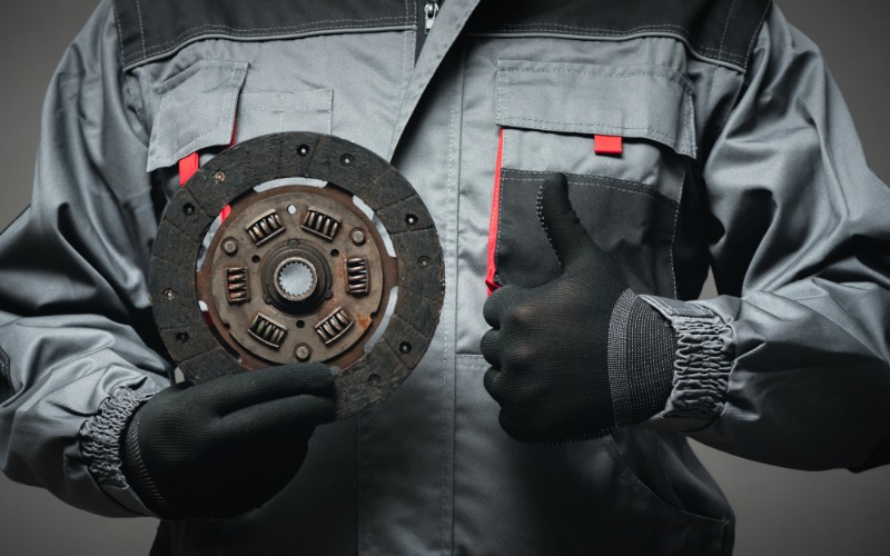 A close-up of the torso of a person with a worker's jacket and black gloves holding up an old car clutch disc.