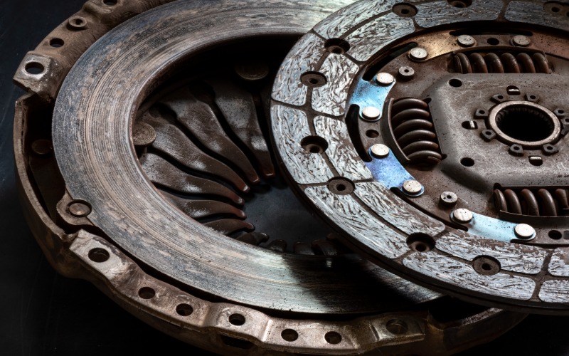 A close-up of a hand with a blue latex glove holding a new car clutch disc next to an elevated vehicle's transmission.