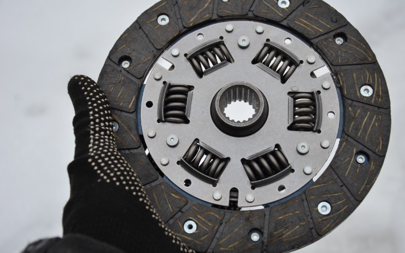 A close-up of a hand with a heavy-duty black glove holding out the clutch disc of a manual transmission assembly.