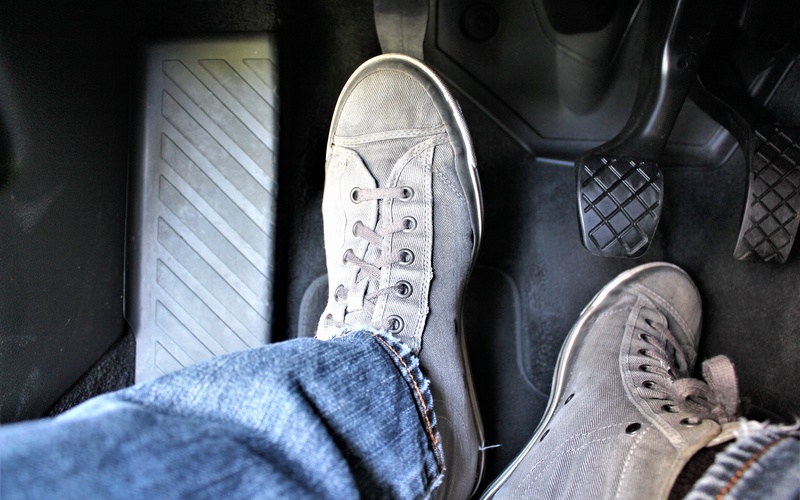 A close-up of the feet of a driver, with tennis shoes and jeans, operating the clutch pedal of a car.