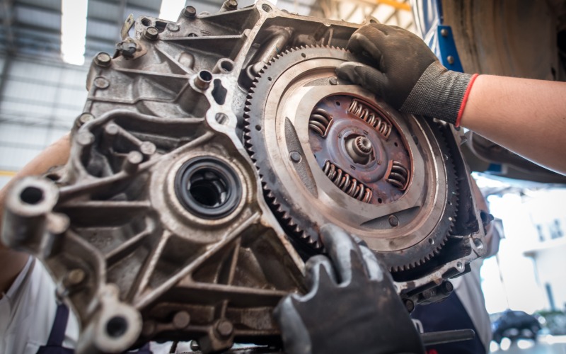 A close-up of a pair of gloved hands touching the exposed automotive flywheel of a disconnected automotive transmission.