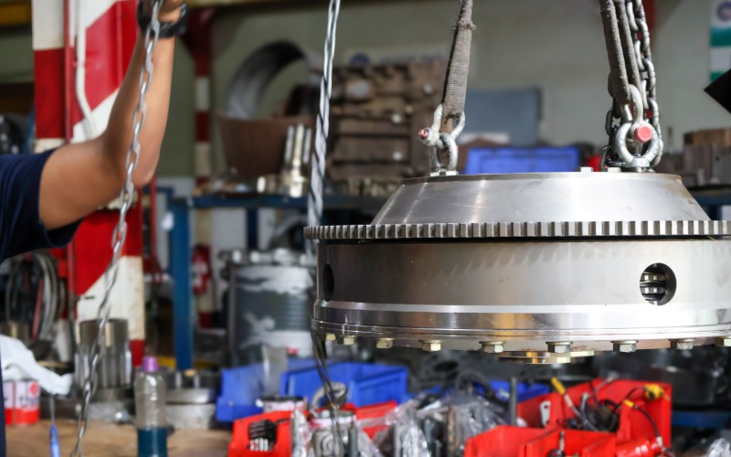 A close-up of the side of a heavy steel automotive flywheel being hoisted by a chain hoist in an auto workshop.