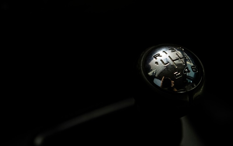 A close-up of the gear shift lever of a six-speed car. The stick shows six gears and reverse on a reflective metal handle.