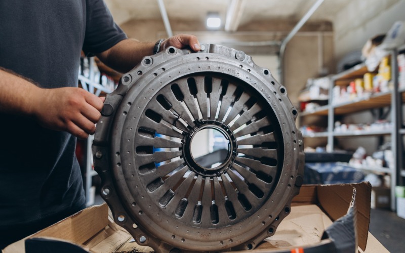 A man in a grey T-shirt holding upright a new automotive clutch pressure plate on top of a box in an auto workshop.