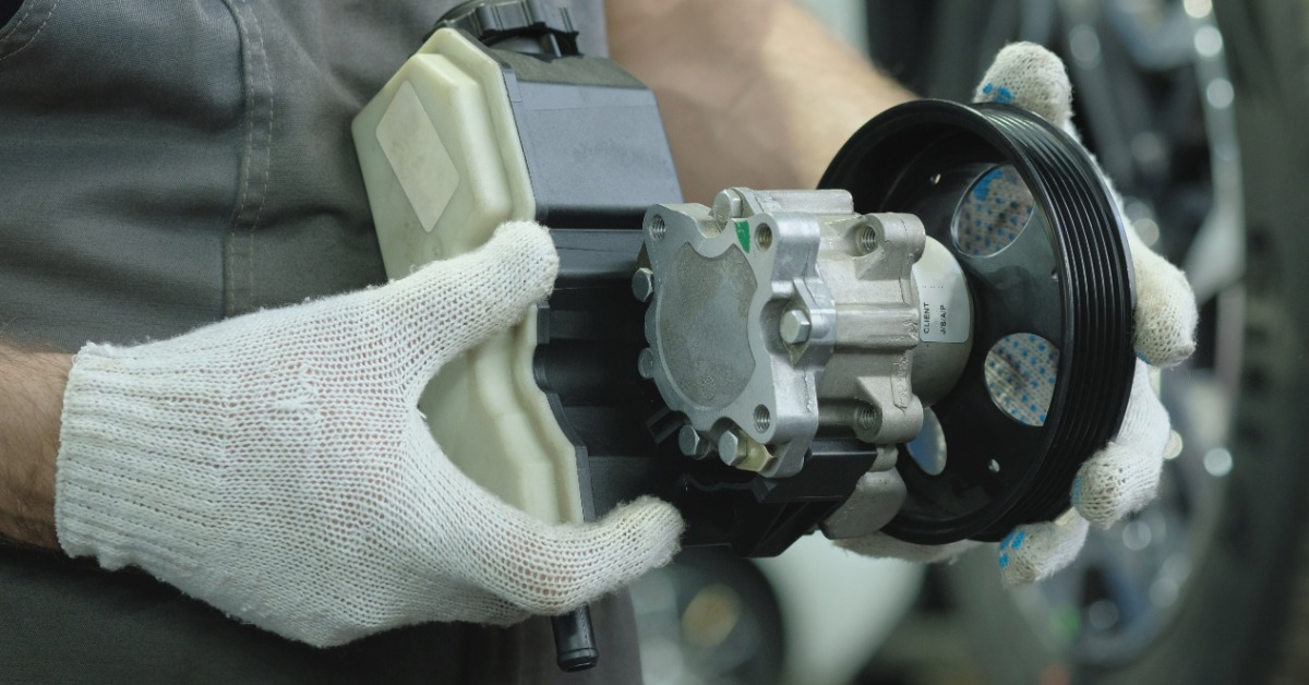 A close-up of a man in a grey apron and white cotton gloves using both hands to hold a new hydraulic pump for a car.