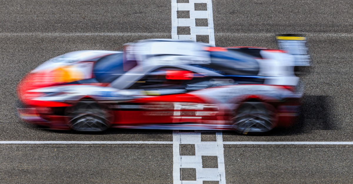 An action shot of a high-speed race car with a white and red livery crossing the checkered finish line.