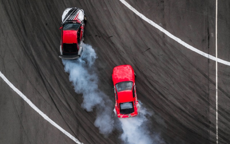 An overhead view of two cars on a racetrack drifting around a corner and creating smoke from their rear tires.