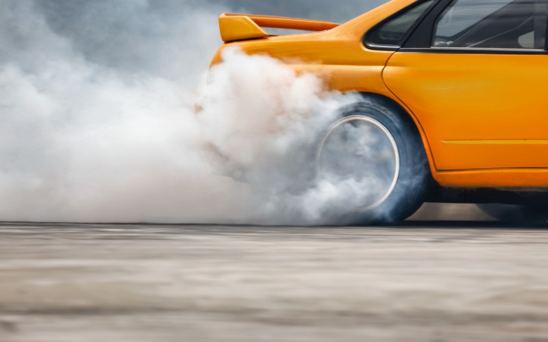 The rear end of a yellow, four-door sports car with a spoiler doing a tire burnout and creating clouds of smoke.