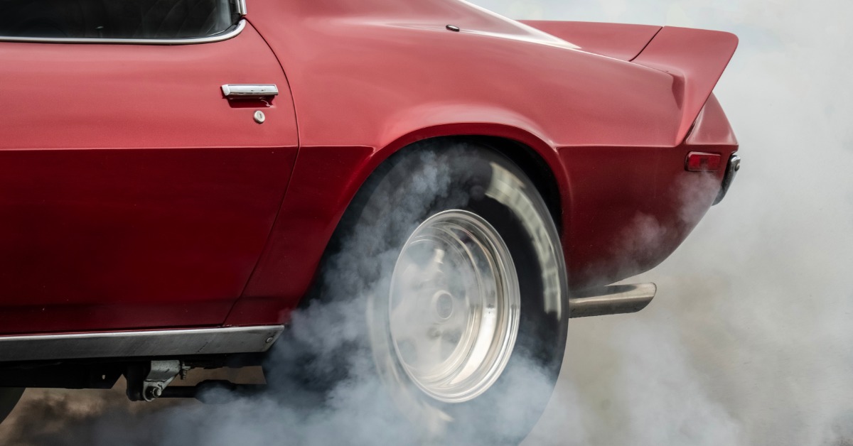 A close-up of the rear tire and end of a red, two-door classic sports car as it burns rubber and produces smoke.