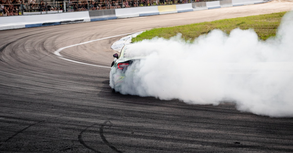 A rear view of a green race car drifting around the corner of a race track and producing large plumes of smoke.