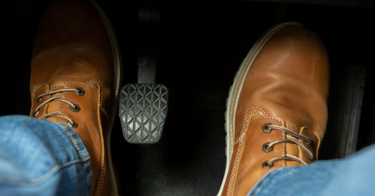 A close-up of the feet of a driver, wearing brown leather boots and blue jeans, operating the gas and clutch pedal.