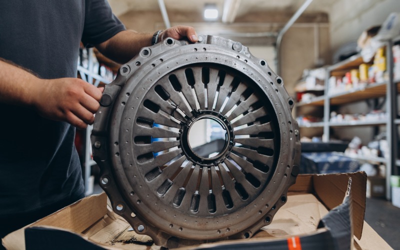 A close-up of a new automotive clutch pressure plate, propped upright by a man in a black T-shirt in an auto shop.