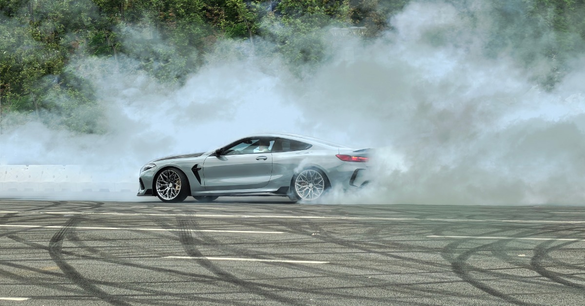 A silver, two-door sports car on a large asphalt area does burnouts as plumes of smoke arise from its rear tires.