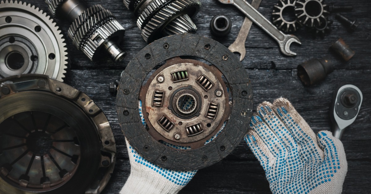 A point of view of a person with white gloves holding an old clutch disc above a table with other clutch parts.