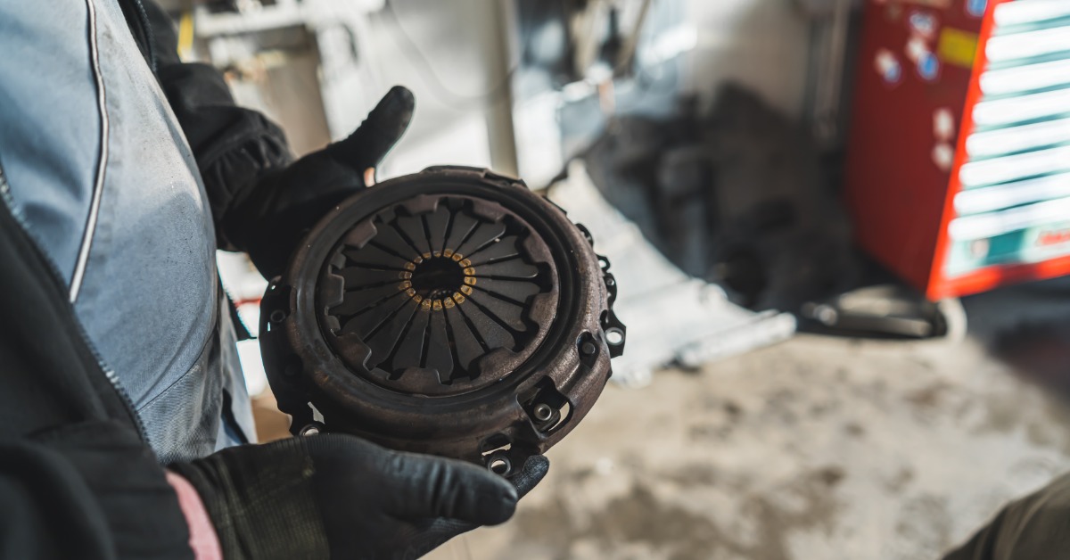 A close-up of a person in overalls with black gloves holding onto the used clutch of a car in a garage.