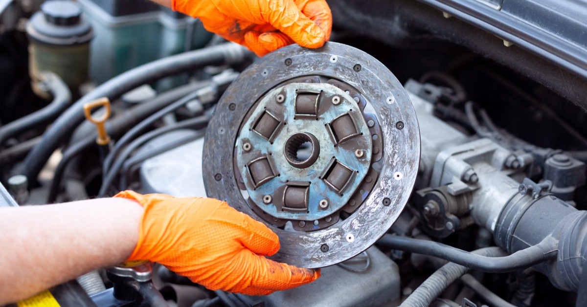 A close-up of a pair of hands with orange latex gloves holding up a broken car clutch disc over the engine bay.