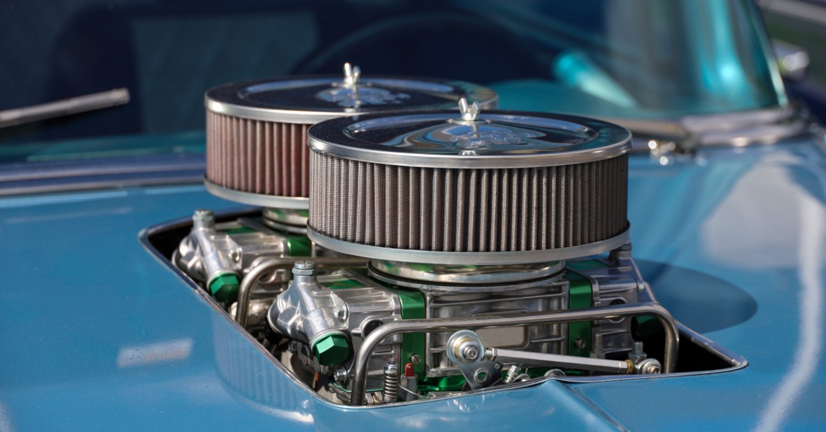 A close-up of the hood of a blue classic car with a cut-out for a dual-quad carburetor setup with chrome air cleaners.