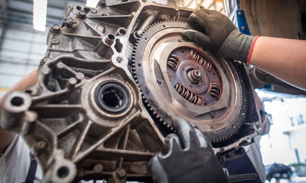 A mechanic’s gloved hands holding up a mounted car engine in an auto shop with the flywheel exposed.