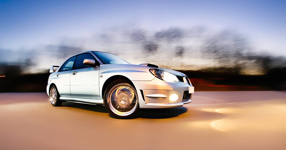 A silver Subaru WRX rally car with chrome wheels and a racing spoiler drifting on an empty road at dusk.